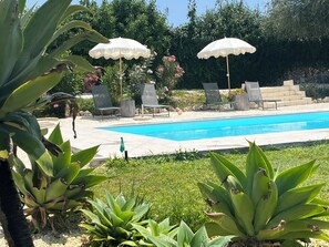 Un espace piscine agrémenté de chaises longues et de parasols, entouré de verdure et bordé d’un mur en pierre.
