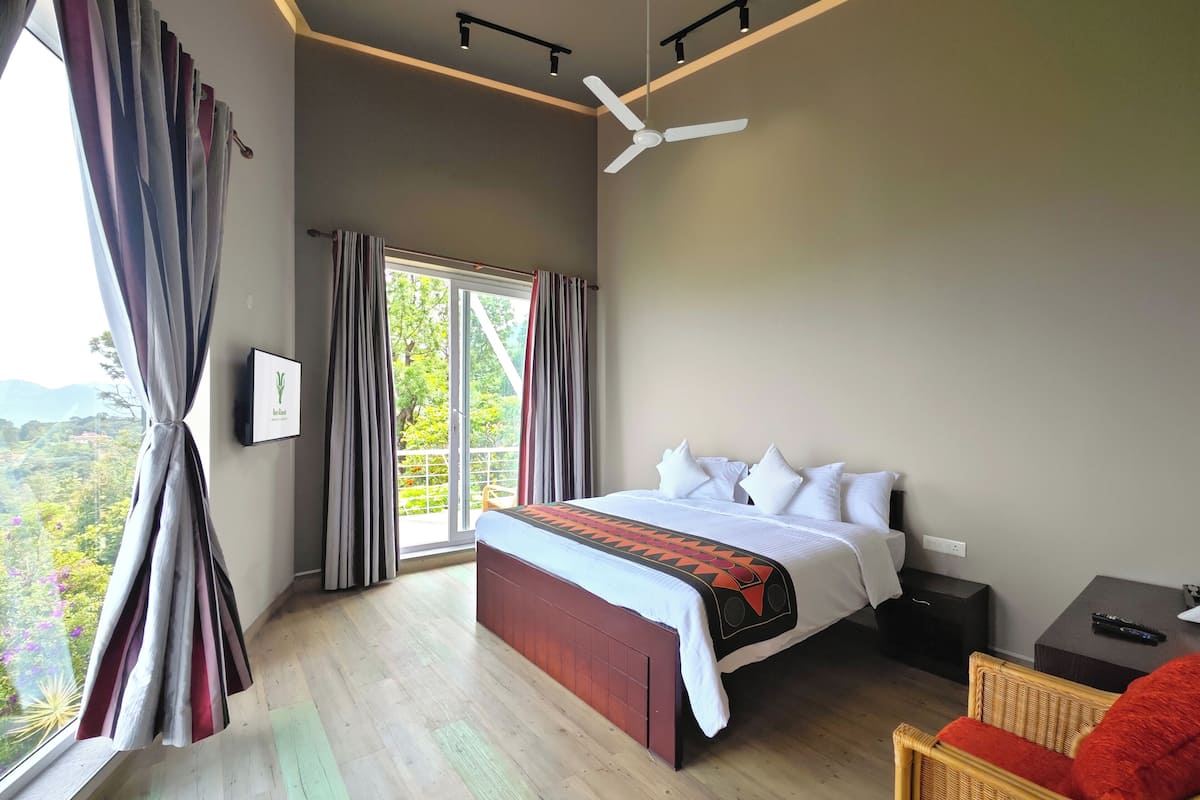 A bedroom with a large bed, a ceiling fan, a window with curtains, and a view of greenery outside.