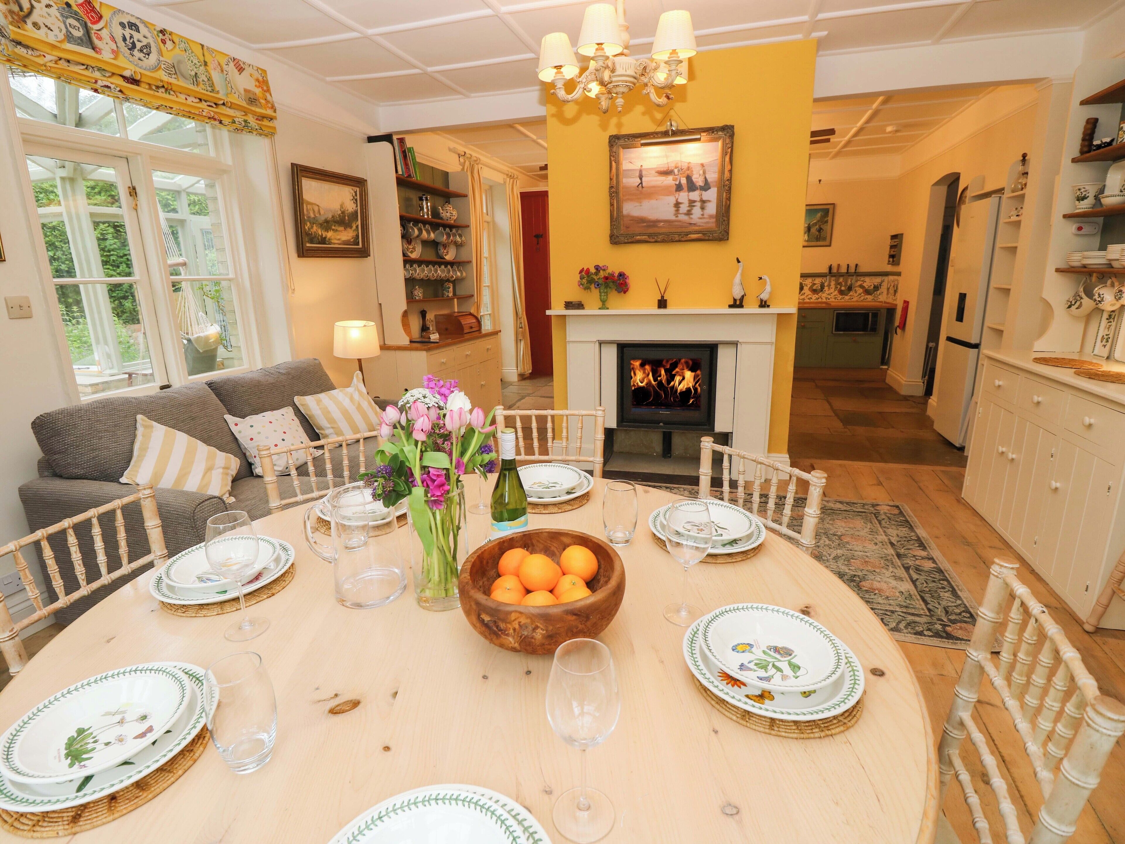 A dining area with a round table set for four, surrounded by chairs. A fireplace is visible in the background, and there is a bookshelf with various items on the wall.