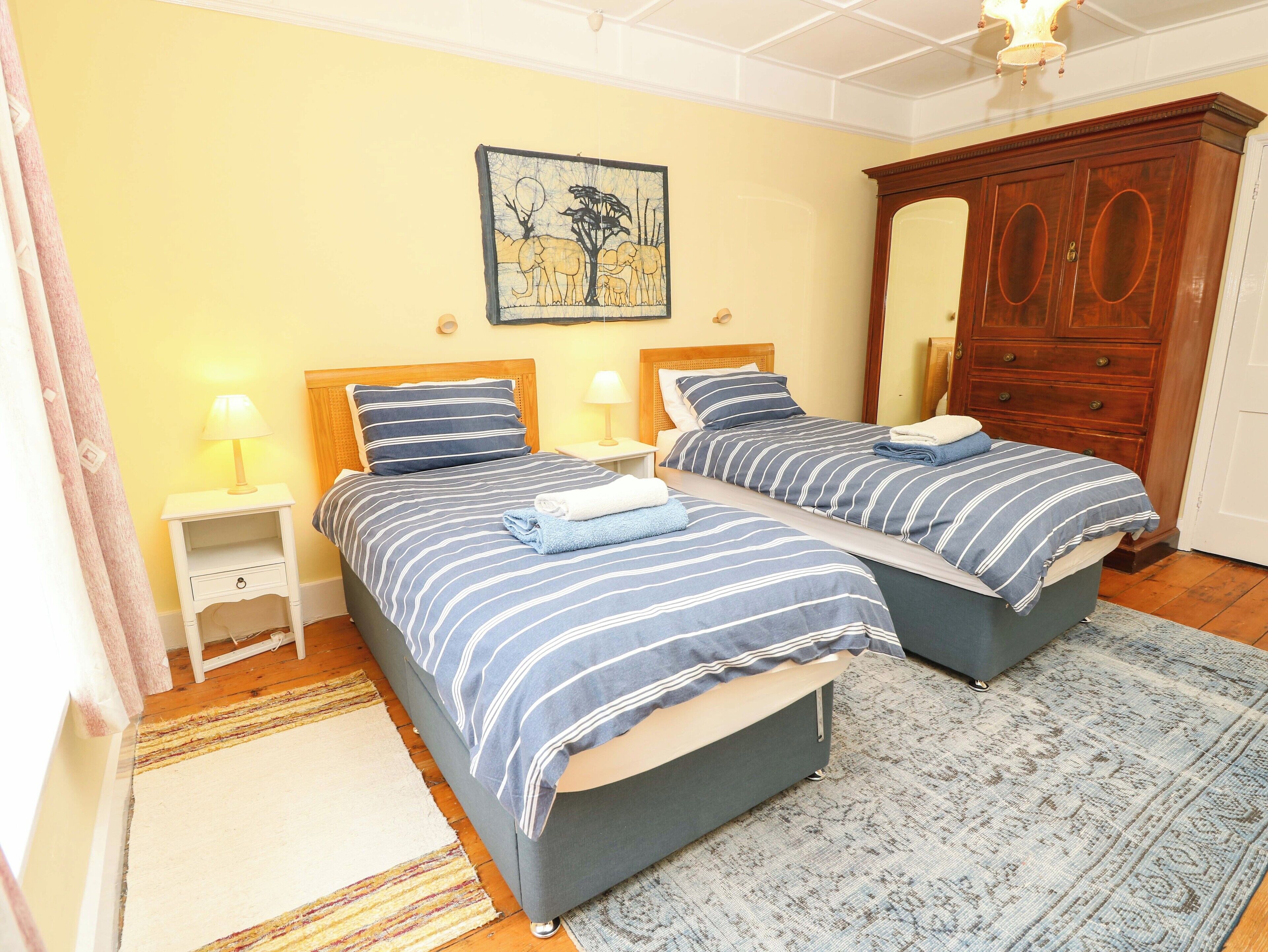 A double room with two beds, each with striped bedding, a wooden wardrobe, a painting on the wall, and a small bedside table with a lamp.