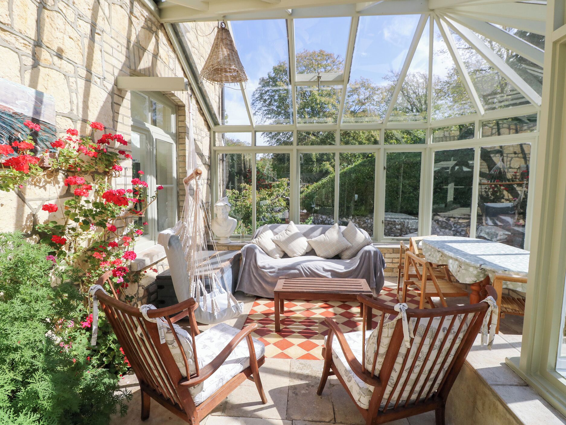 A conservatory with a stone wall, a cushioned bench, wooden chairs, a table, and a hanging light fixture.