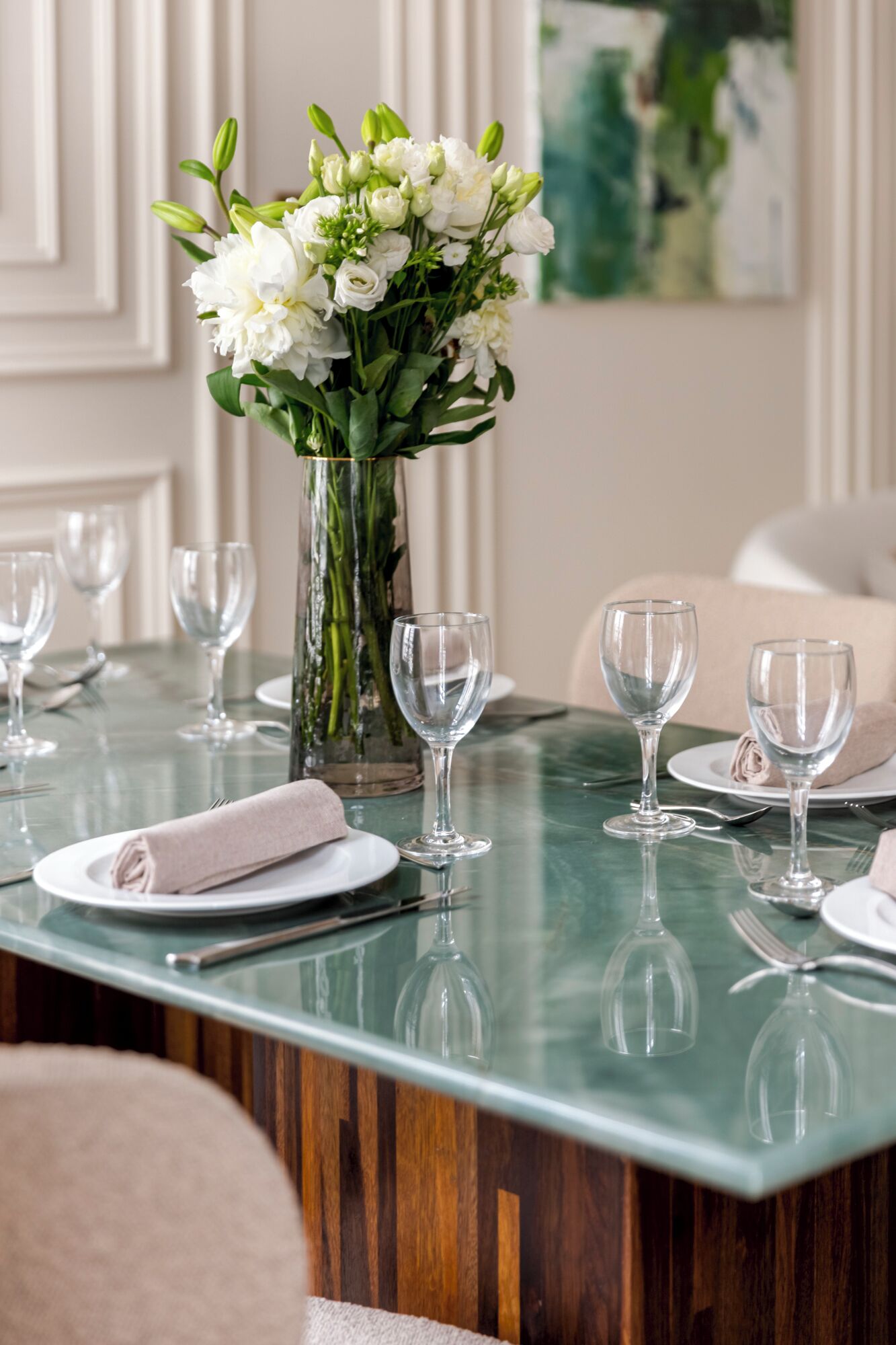 Uma mesa de jantar posta com pratos brancos, copos de vinho transparentes e um vaso de flores brancas.