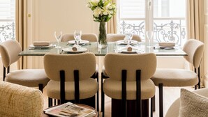 A dining area with a round table set for six, surrounded by six chairs. There is a vase with flowers on the table, and a magazine on a side table. - Merveil Luxury Suites - Marais - Caire I-II (Paris)