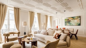 A spacious living room with a sofa, armchairs, and ottomans, a dining area with chairs and a table, and a large abstract painting on the wall. - Merveil Luxury Suites - Marais - Caire I-II (Paris)