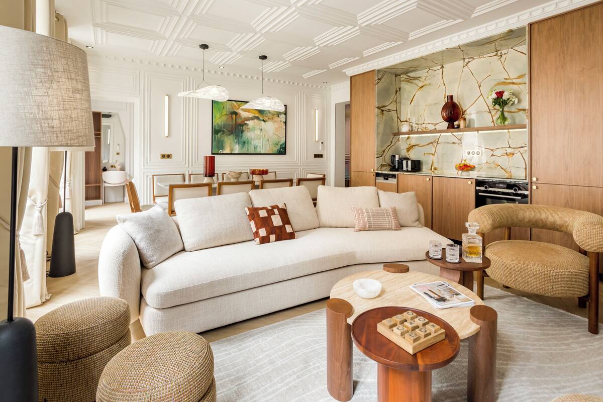 Un salon moderne avec un canapé, une table basse et des fauteuils en osier. Un tableau orne le mur, et on aperçoit un coin cuisine au fond de la pièce.