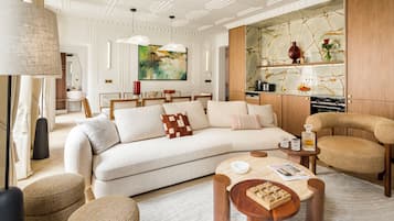A modern living room with a sofa, coffee table, and wicker chairs. There is a painting on the wall and a kitchen area in the background.