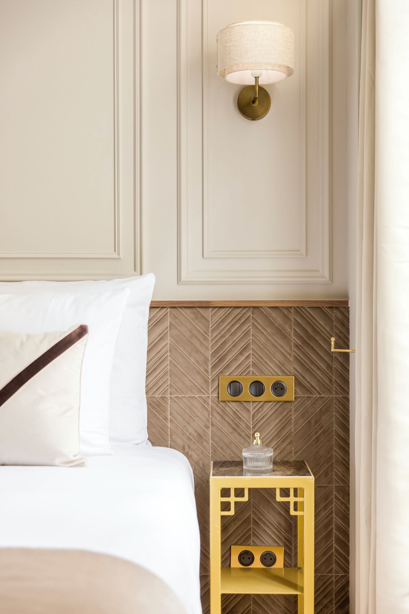 Une chambre d’hôtel avec un lit, une lampe fixée au mur, une table de chevet sur laquelle se trouve une carafe en verre, et un mur orné d’un motif en chevrons.