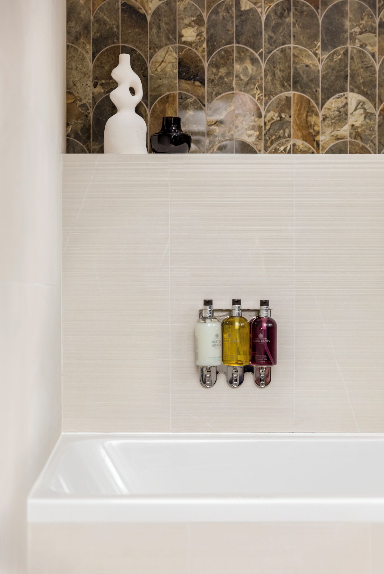 A bathroom with a tiled wall, a white vase, and three bottles of toiletries.