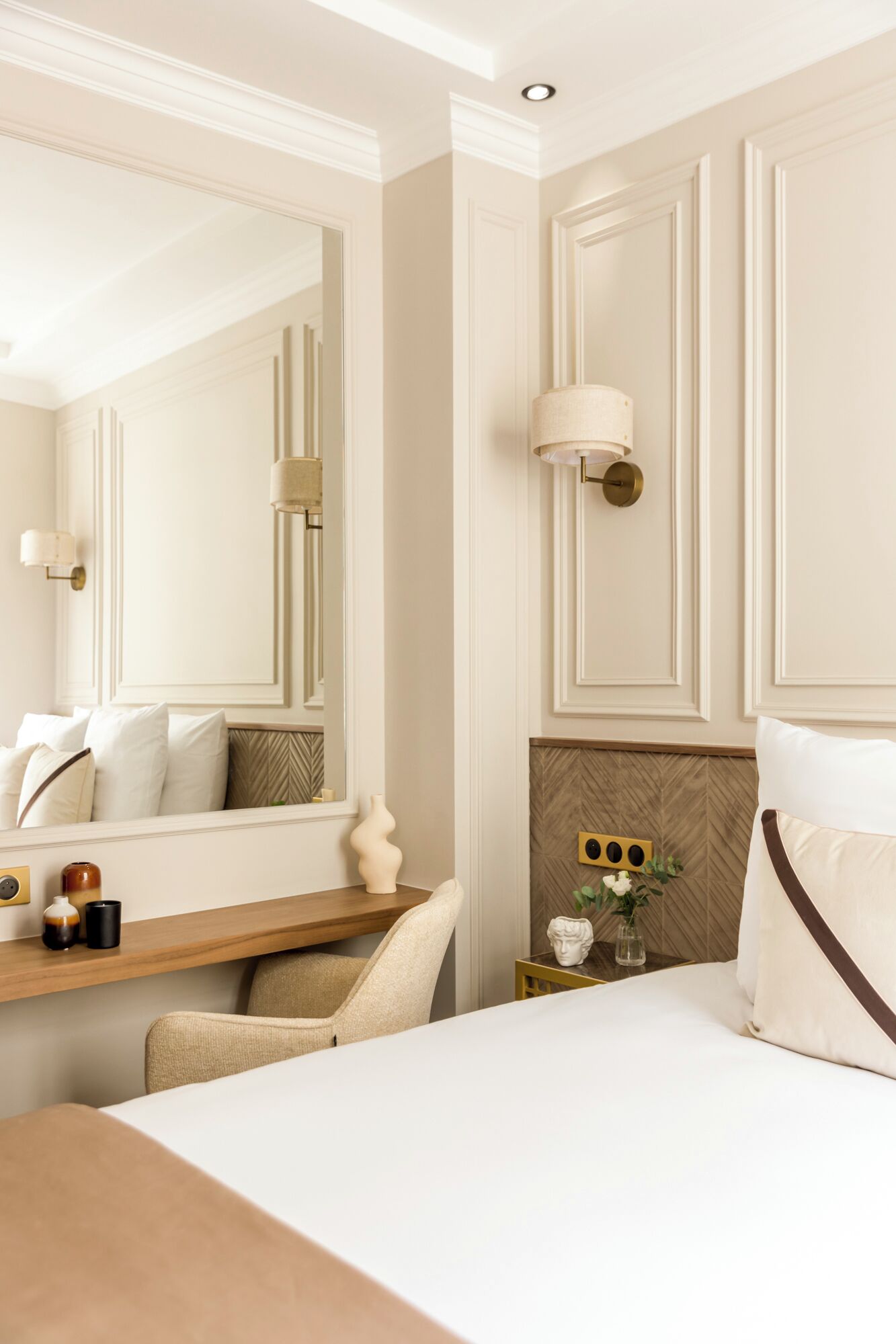 Habitación de hotel con una cama grande, una mesita de noche con lámpara, un espejo y una pequeña mesa auxiliar con objetos decorativos.