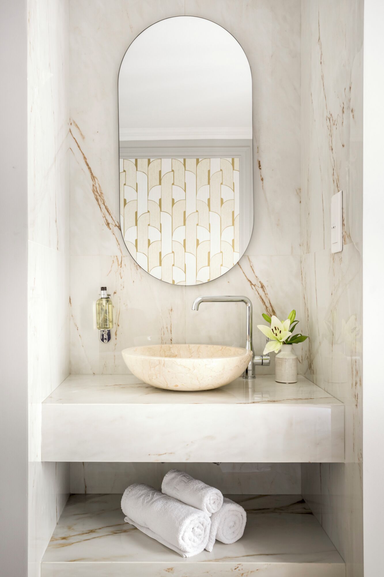 Un bagno con un lavabo in marmo, uno specchio rotondo, un dispenser di sapone a parete e un vaso con un fiore.