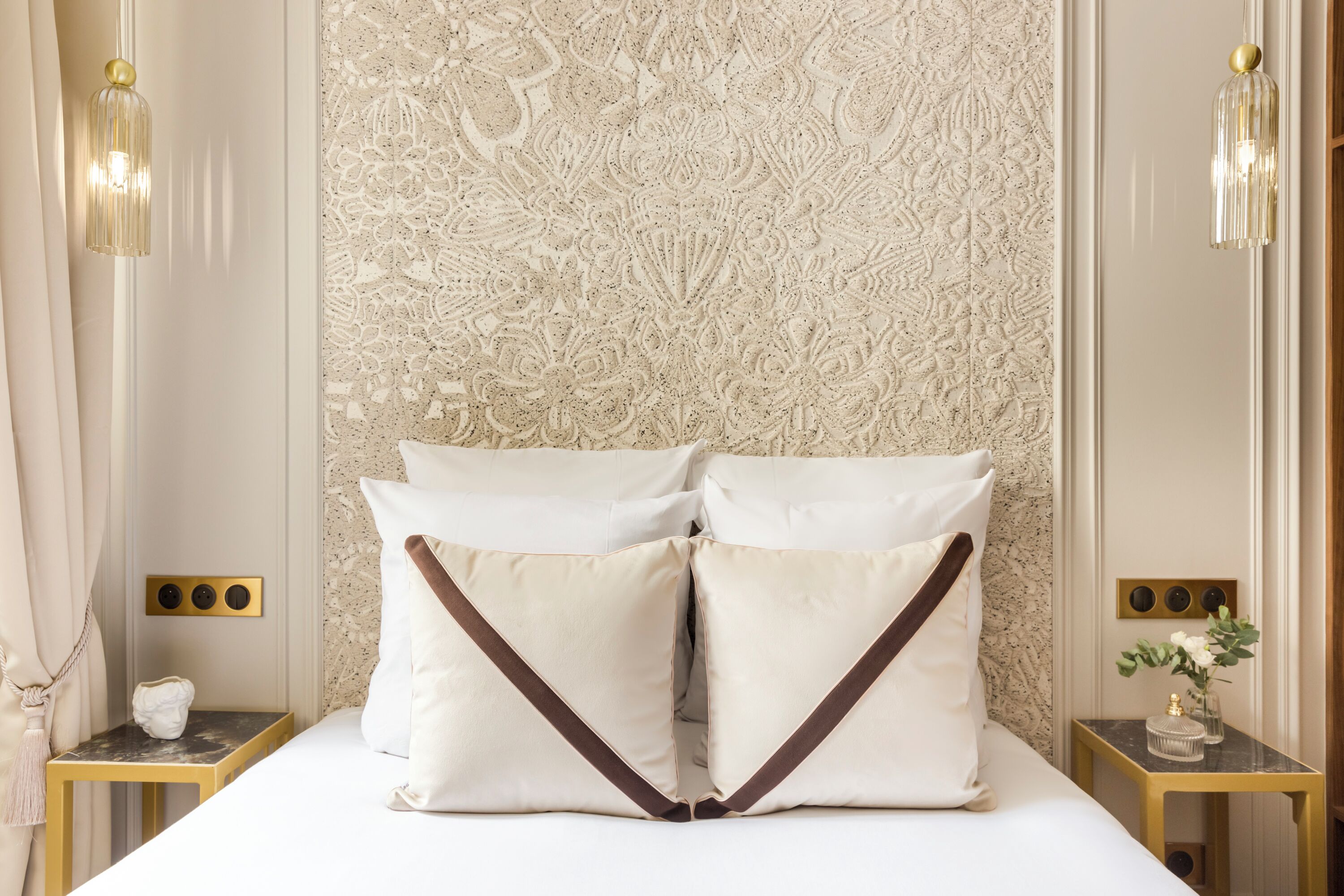 Een net opgemaakt bed met witte lakens en twee kussens met bruine randen, een wand met een marmerpatroon erachter en twee nachtkastjes met lampen.
