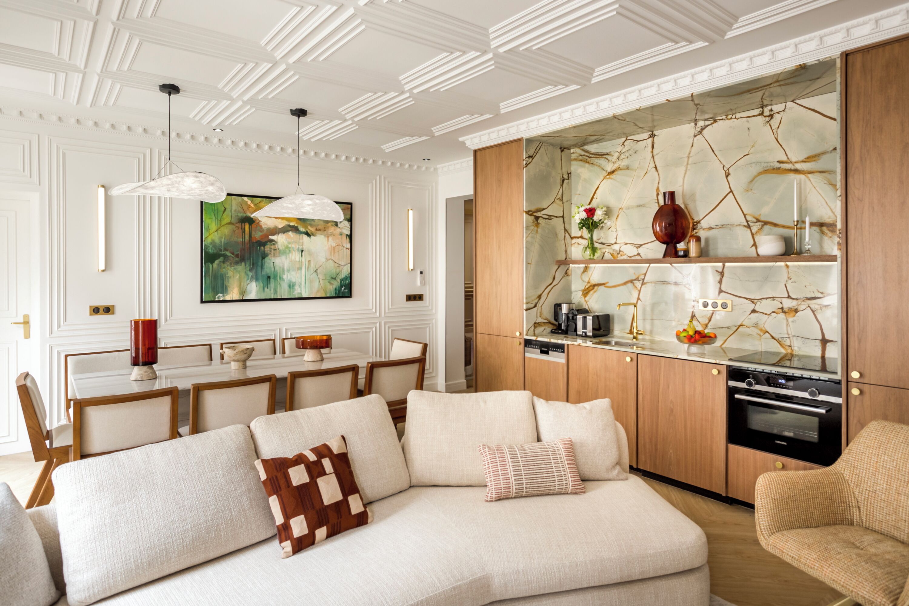 A modern living room with a sectional sofa, a dining area with a table and chairs, and a kitchen with marble backsplash and wooden cabinets.
