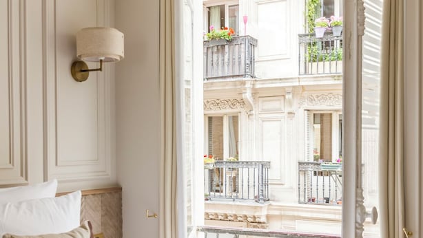A hotel room with a bed, a nightstand, and a view of a balcony with ornate railings and potted plants. - Merveil Luxury Suites - Marais - Caire I-II (Paris)