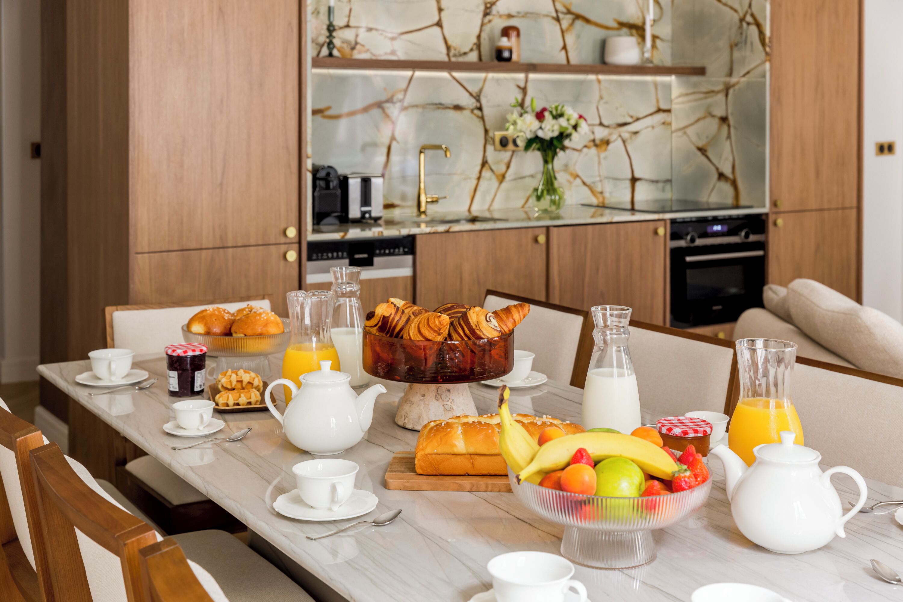 A breakfast table with pastries, fruits, and beverages, a kitchen in the background, and a modern interior design.