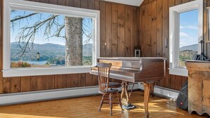 A room with wooden walls, a piano, a chair, and a large window offering a view of a tree and mountains.