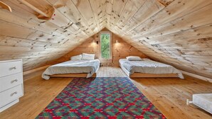 A small, cozy attic bedroom with two single beds, a colorful rug, and a window with a view of greenery.