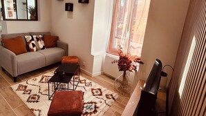 A living room with a sofa, coffee table, ottoman, and a TV. There is a window with curtains, a plant, and a vase with flowers. - Cosy house (Pontarmé)