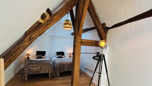 A loft bedroom with exposed wooden beams, a wooden floor, two beds, a tripod, and a hanging light fixture. - Cosy house (Pontarmé)