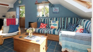 A cozy loft-style living room with a sloped ceiling, a sofa with striped cushions, a wicker chair, and a wooden coffee table. - Borth Arian Boat House (Holyhead)