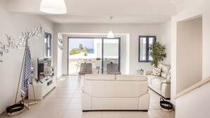 A modern living room with a white sofa, a TV, and a dining area with chairs and a table. There is a balcony with a view of the outside. - Protaras Seaside Villa Protamare 32 (Paralimni)