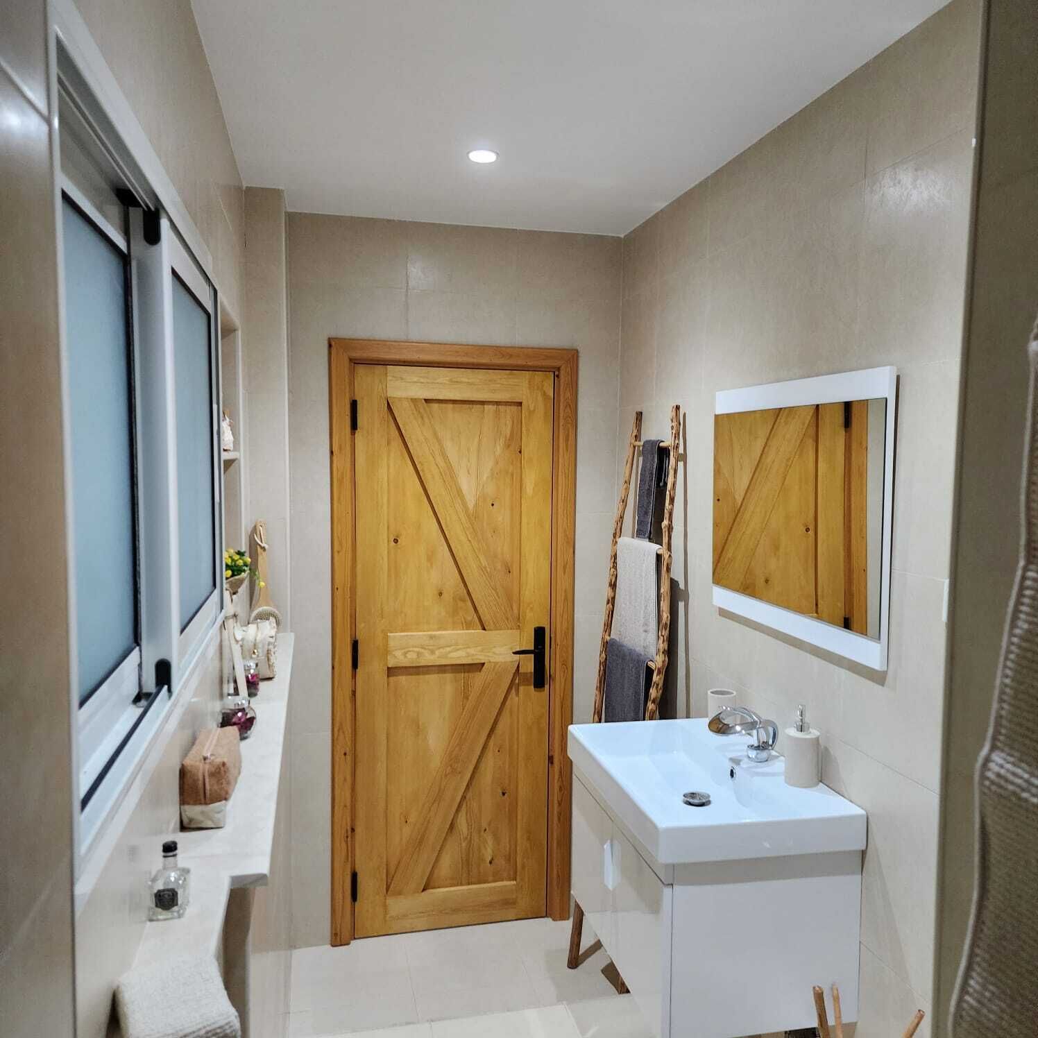 Une salle de bain avec une porte en bois, un lavabo blanc, un miroir et une fenêtre.