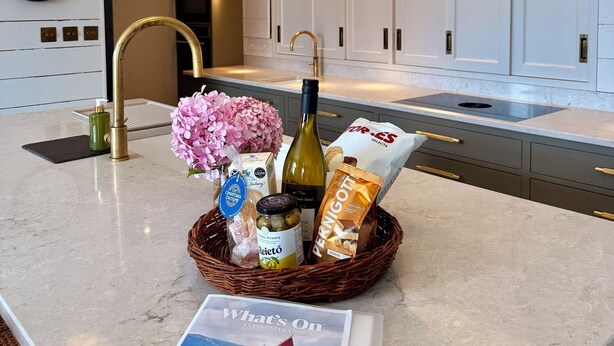 A modern kitchen with white cabinets, a marble countertop, and pendant lighting. A basket with food items and a wine bottle is on the counter. - The Lodge @Sunnybank House - unique country lodge overlooking Clifden Town (Clifden)