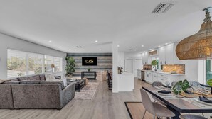Una sala moderna con un sofá gris, un comedor con mesa dispuesta para dos personas, y una cocina con armarios blancos y una lámpara colgante.