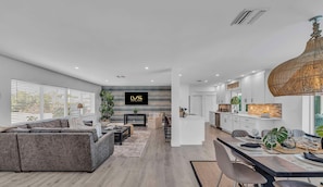 Un salon moderne avec un canapé gris, un coin repas avec une table dressée pour deux personnes et une cuisine dotée de meubles blancs et d’un luminaire suspendu.