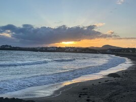 Escena costera con olas, una playa y edificios al fondo.