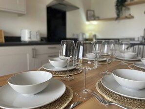 Mesa de comedor dispuesta con cuencos blancos, copas de vino y cubiertos, con una cocina al fondo.