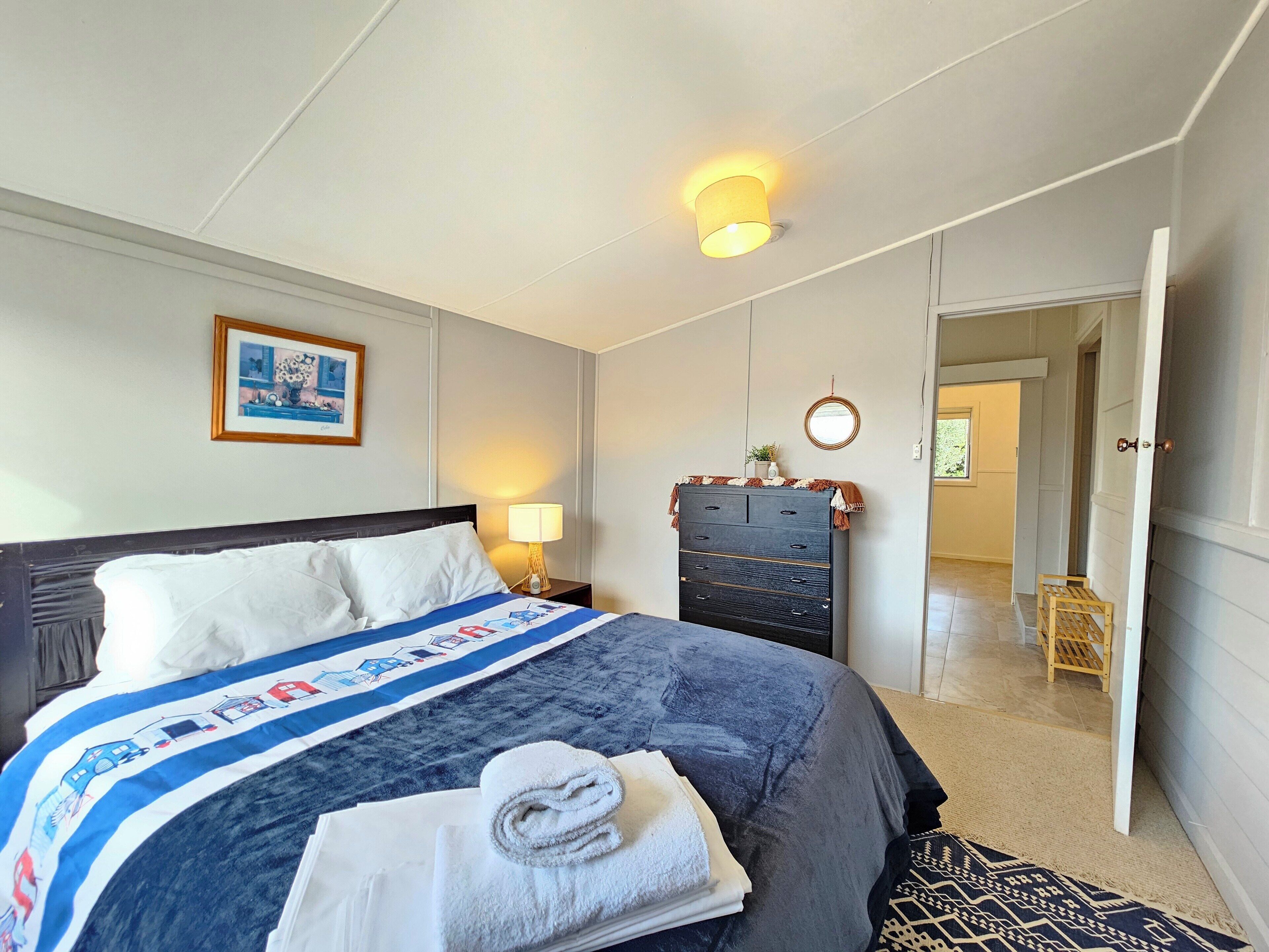 A bedroom with a bed, a dresser, a nightstand, a lamp, a framed picture, and a door leading to another room.