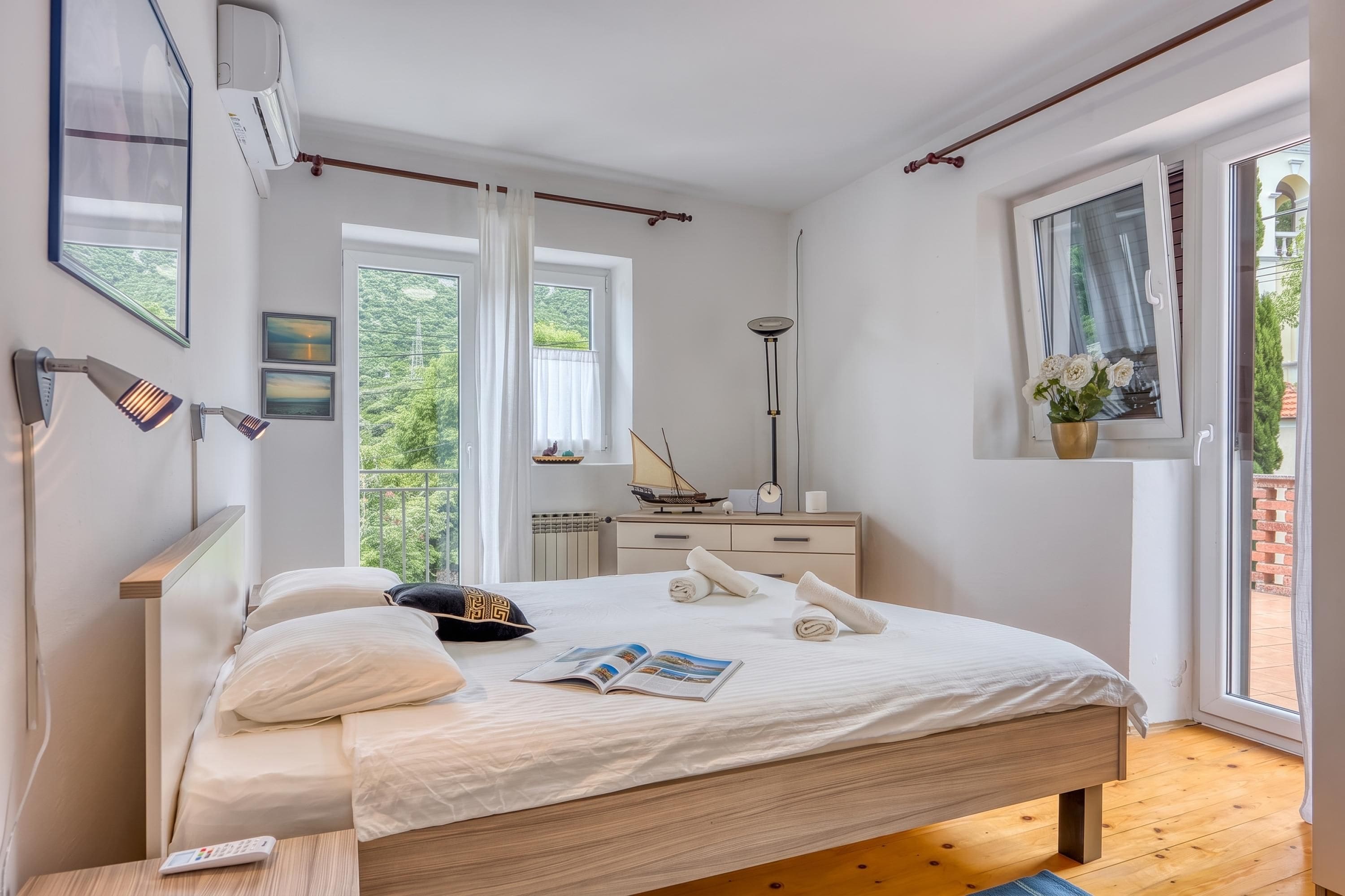 Ein modernes Schlafzimmer mit einem großen Bett, Nachttischlampen, einem Nachttisch, einem Fenster mit Blick auf Grünflächen und einem kleinen Beistelltisch mit einem Blumenstrauß.