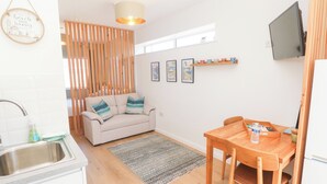 A compact kitchen with a wooden dining table, a sofa with cushions, a small rug, and a TV mounted on the wall. - Carn Brea (Redruth)