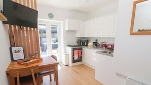 A modern kitchen with white cabinets, a black stove, and a red toaster. There is a wooden dining table with a chair and a framed welcome sign. - Carn Brea (Redruth)