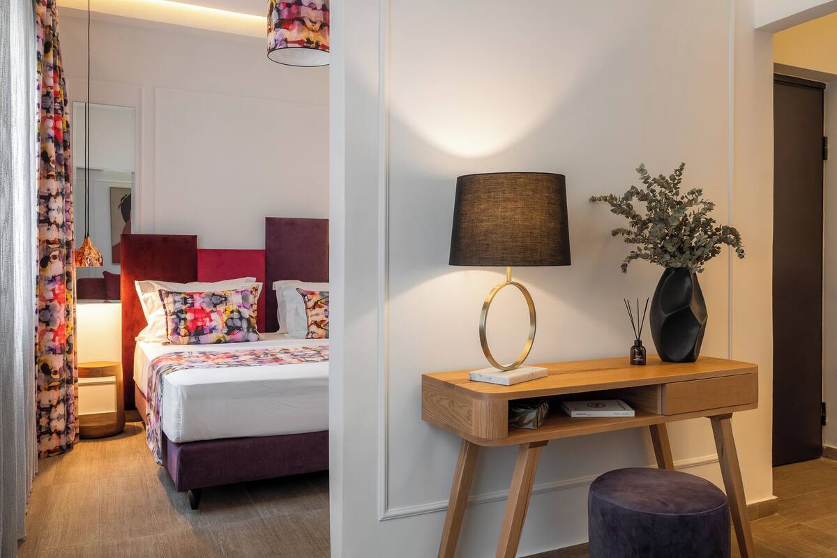 A hotel room with a bed, a desk with a lamp, a vase with flowers, and a mirror.