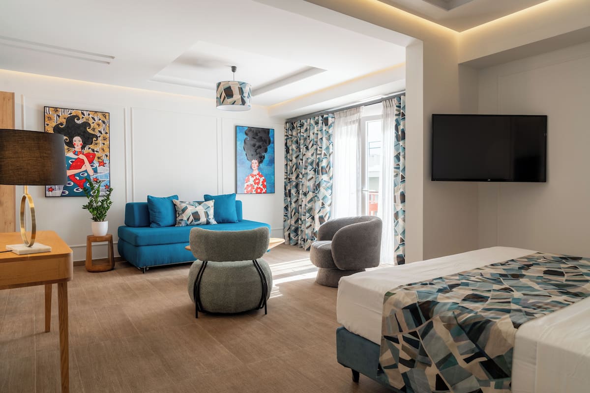 A modern hotel room with a large bed, a flat-screen TV, a blue sofa, a grey armchair, and a painting on the wall.