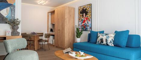 A modern living room with a blue sofa, a wooden coffee table with pastries, and a dining area with a wooden table and chairs.