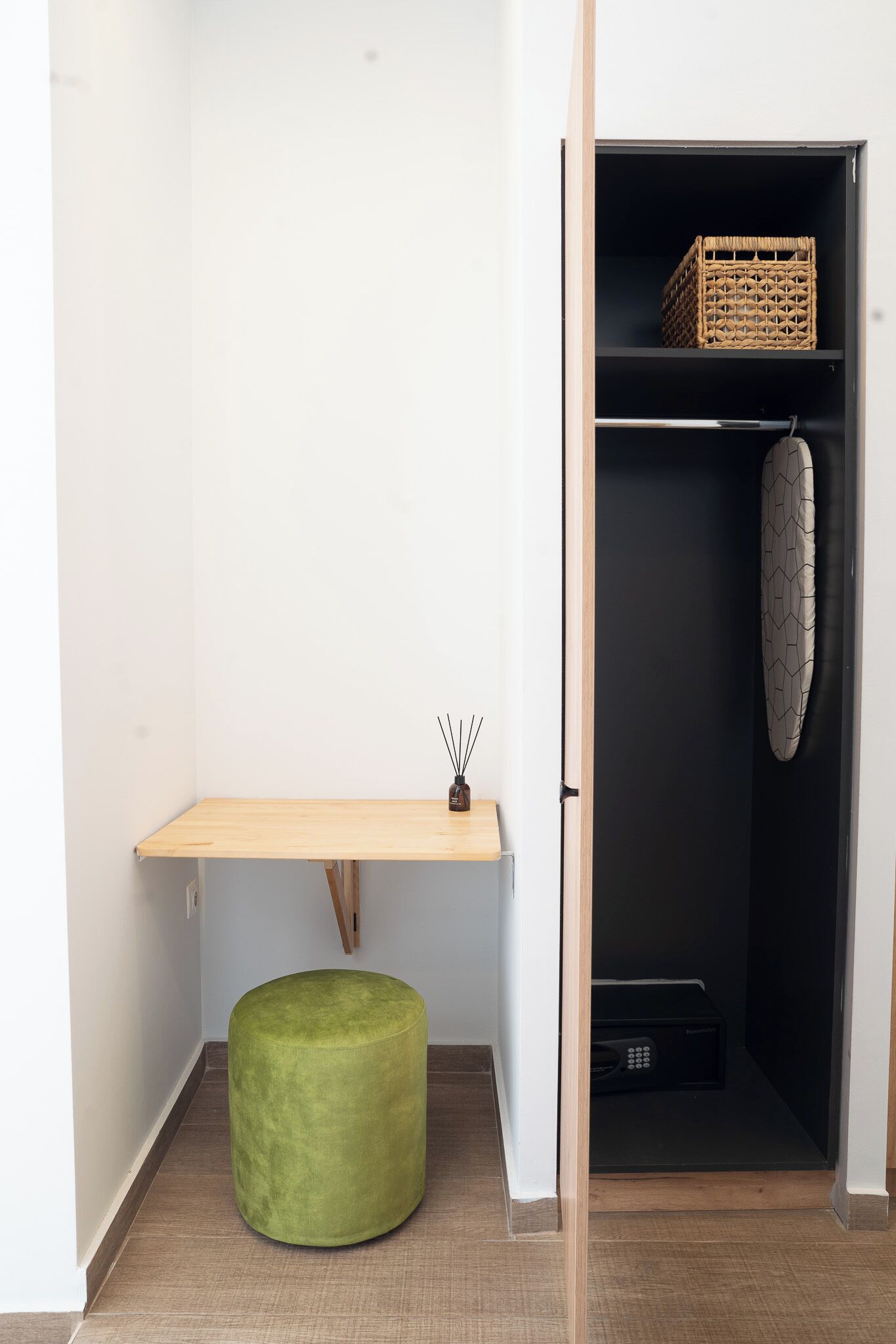 A corner with a wooden bench, a green ottoman, a wicker basket on a shelf, and a black door with a patterned item hanging on it.