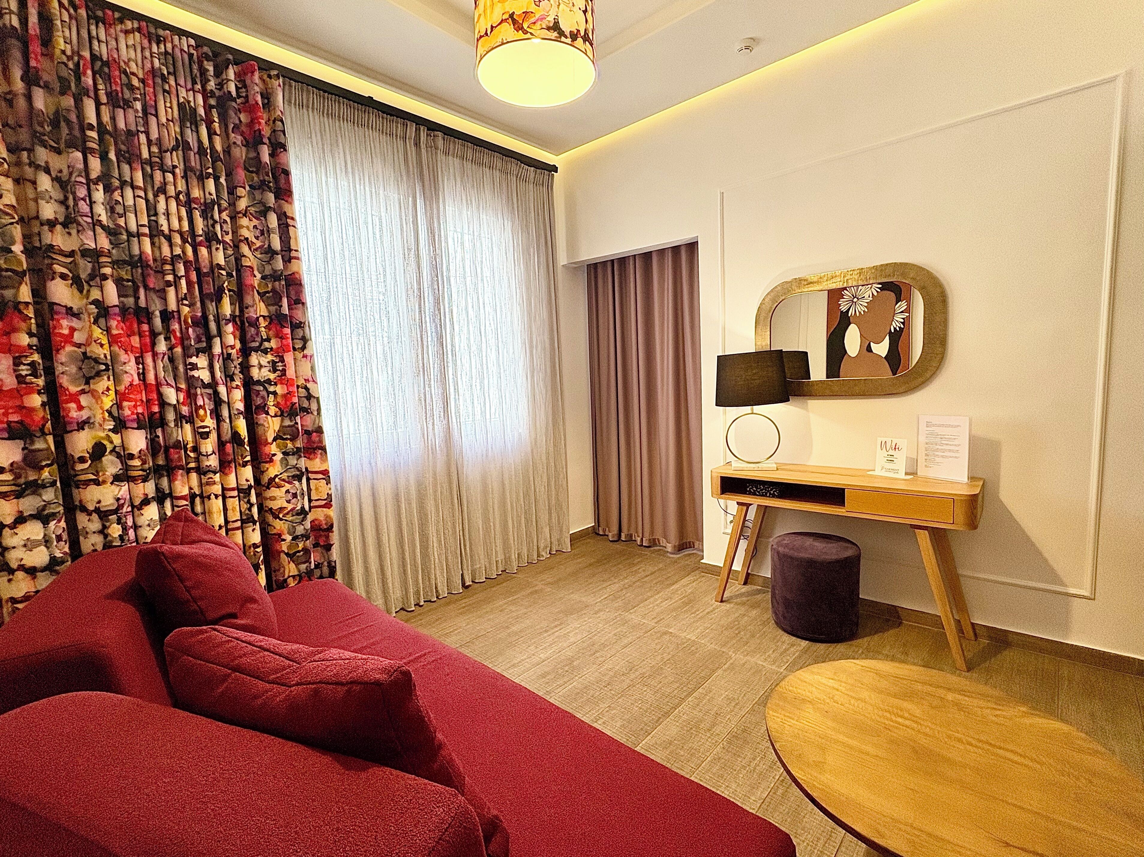 A hotel room with a red sofa, a wooden desk with a lamp, a mirror, and a small round table.