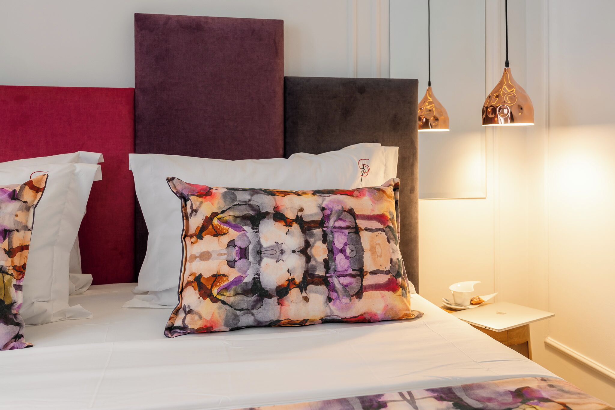 Uma cama bem arrumada com almofadas estampadas, cabeceira vermelha e luminárias pendentes.