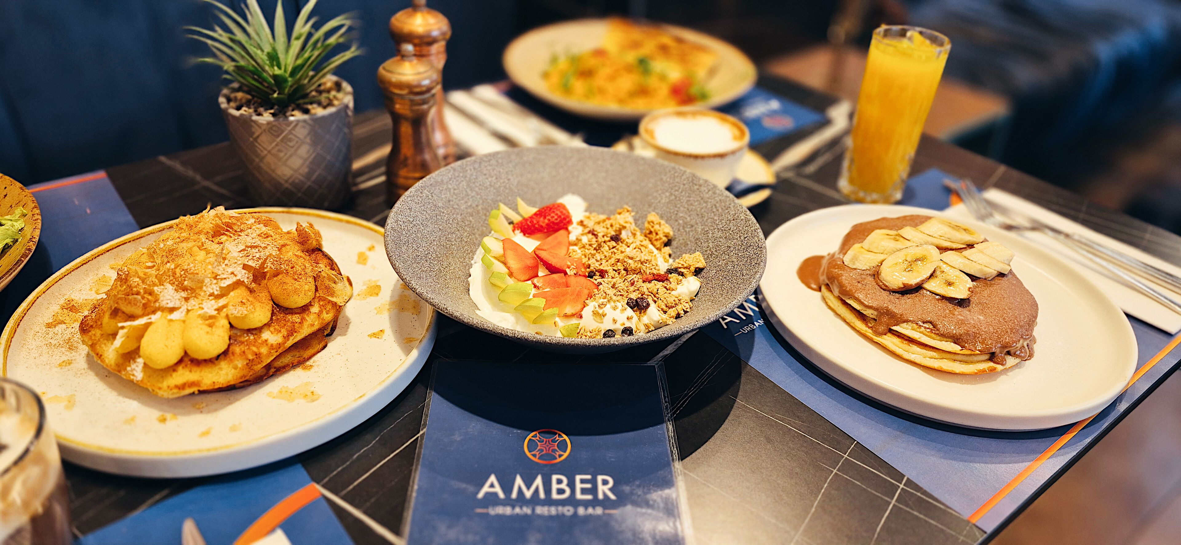 A table with breakfast dishes, a plant, a salt shaker, and a menu titled "AMBER URBAN RESTO BAR".