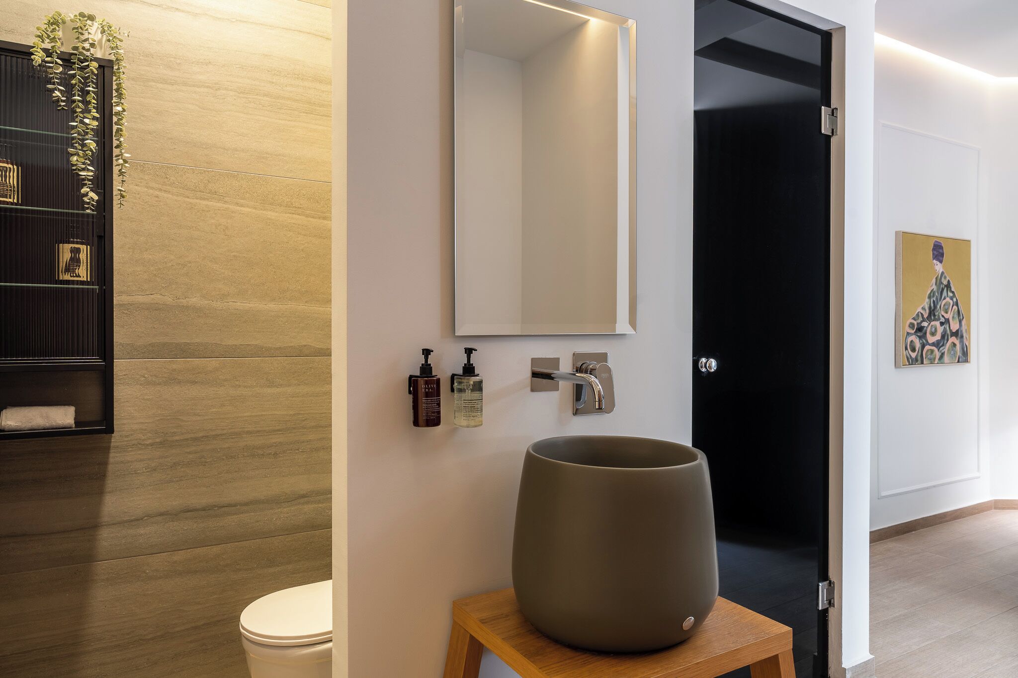 A modern bathroom with a large mirror, a sink, and a toilet. There is a wooden shelf with decorative items and a potted plant.