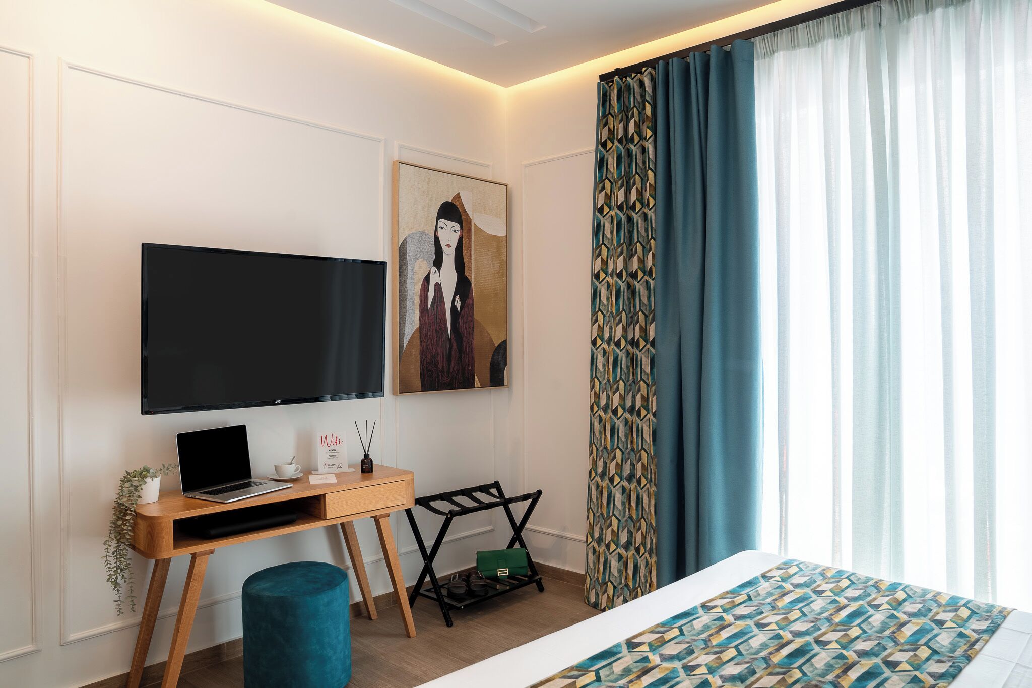 Ein modernes Hotelzimmer mit einem an der Wand befestigten Flachbildfernseher, einem Holztisch mit einem Laptop, einer kleinen Pflanze und einem dekorativen Gegenstand. Ein Bett mit gemustertem Überwurf und ein Fenster mit durchsichtigen Vorhängen sind ebenfalls vorhanden.