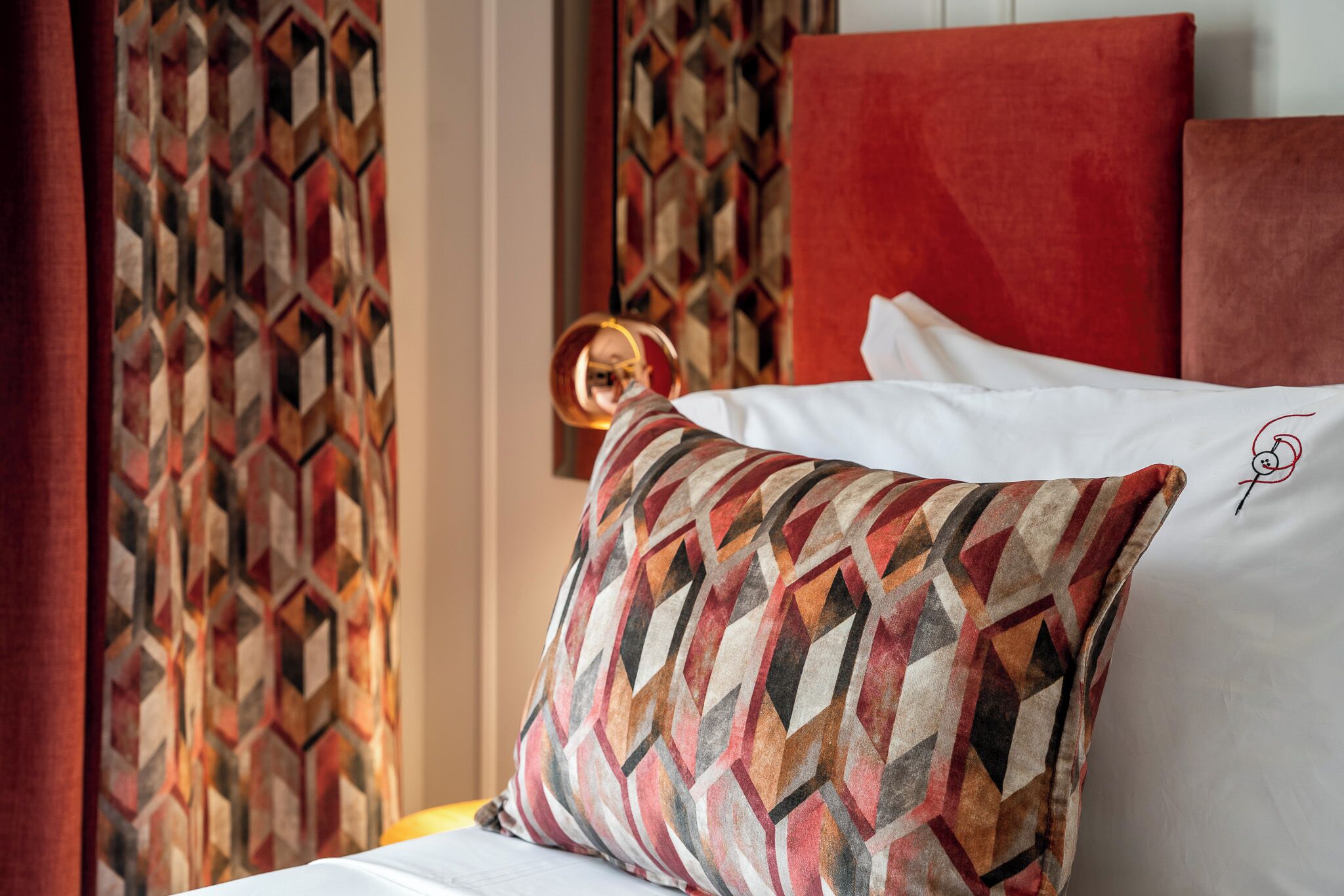 A bed with a patterned pillow and a plain pillow, a red headboard, and a geometric patterned curtain.