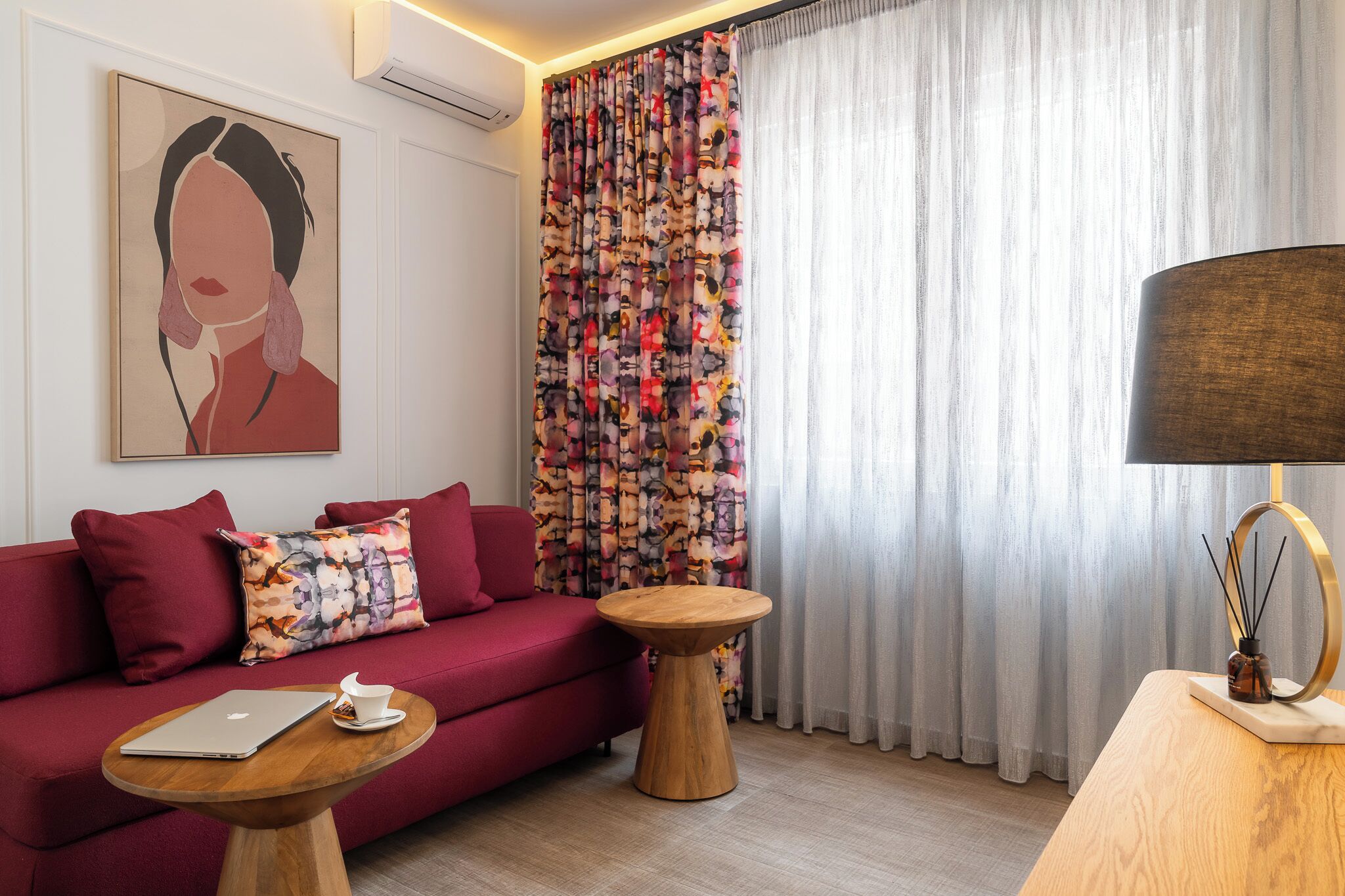 A modern living room with a red sofa, a wooden coffee table, and a small side table. A laptop and a cup are on the coffee table. The room has a large window with sheer curtains and a colorful curtain on the other side.