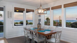 Un espace repas avec une table en bois et des chaises blanches, de grandes fenêtres offrant une vue sur l’océan et un luminaire suspendu.
