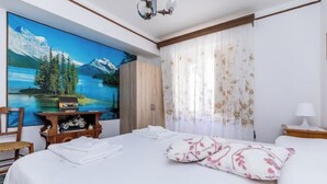 A hotel room with a large bed, a painting of a mountain landscape, a wooden wardrobe, a desk with a lamp, and a chair. - Maria Catena House (Castiglione di Sicilia)