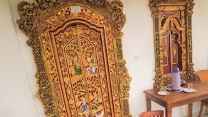 A dining area with a wooden table and chairs, a decorative door with intricate carvings, and a small trash bin. - Kabera Bungalow Ubud (Ubud)