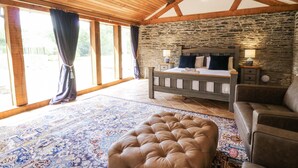 Un dormitorio con pared de piedra, techo de madera, cama con almohadas, mesita de noche, lámpara y un otomano grande.
