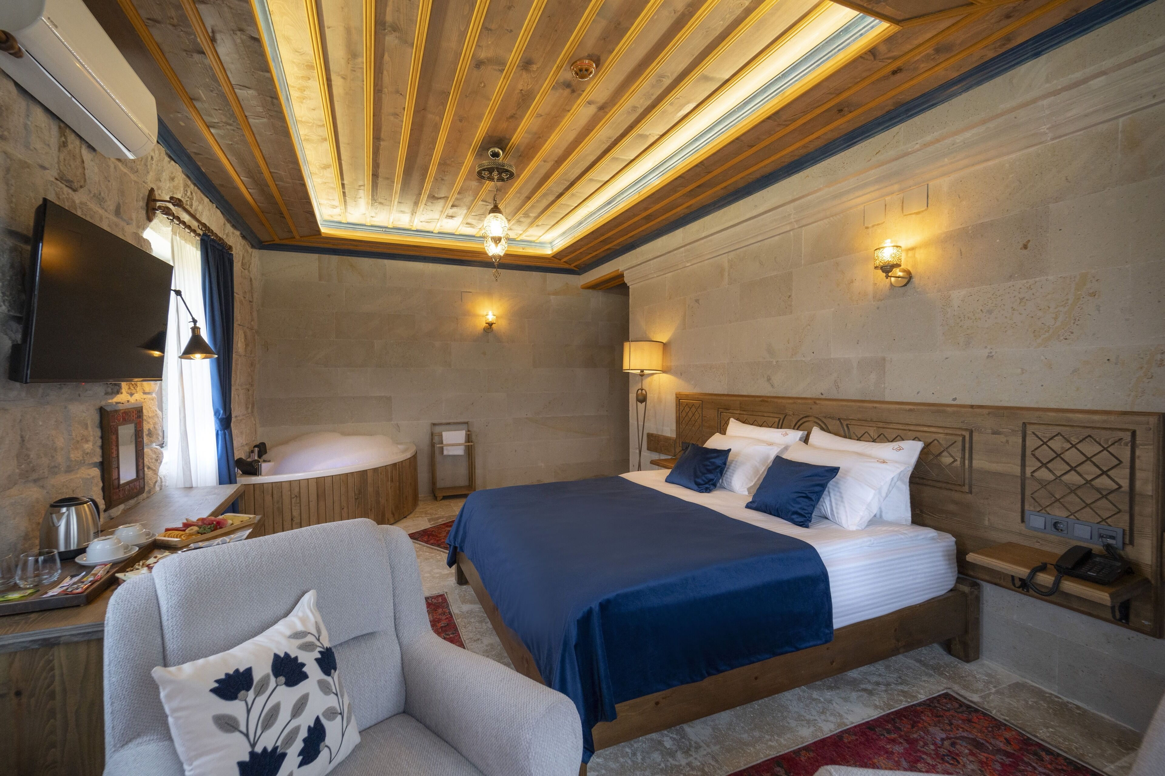 A bedroom with a wooden ceiling, a large bed with blue bedding, a bedside table with a lamp, and a flat-screen TV mounted on the wall.